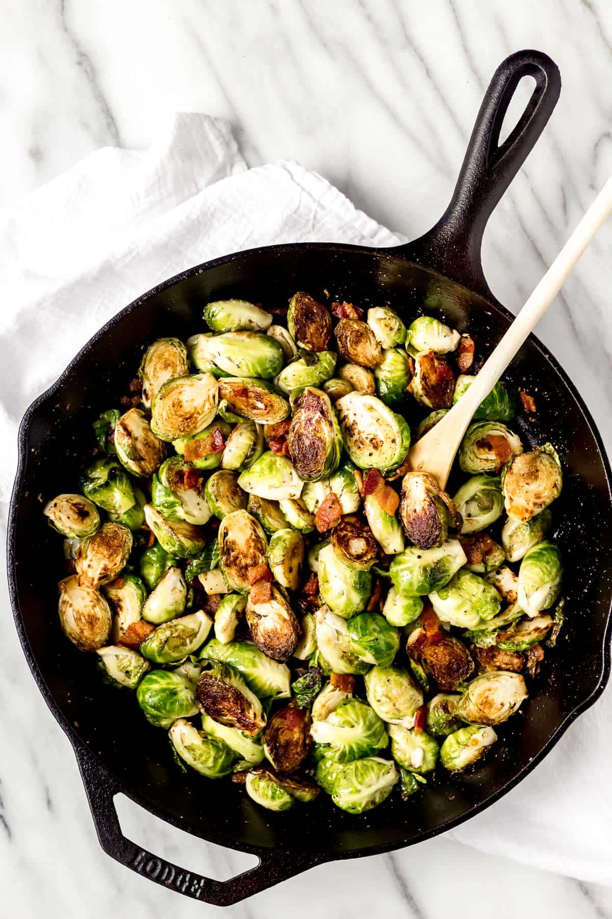 Overhead of brussels sprouts and bacon in a cast iron skillet with a wood spoon over a marble background