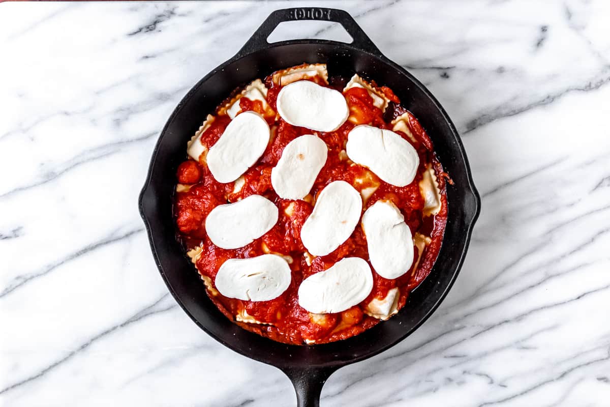 Frozen ravioli topped with sauce and slices of mozzarella cheese in a cast iron skillet