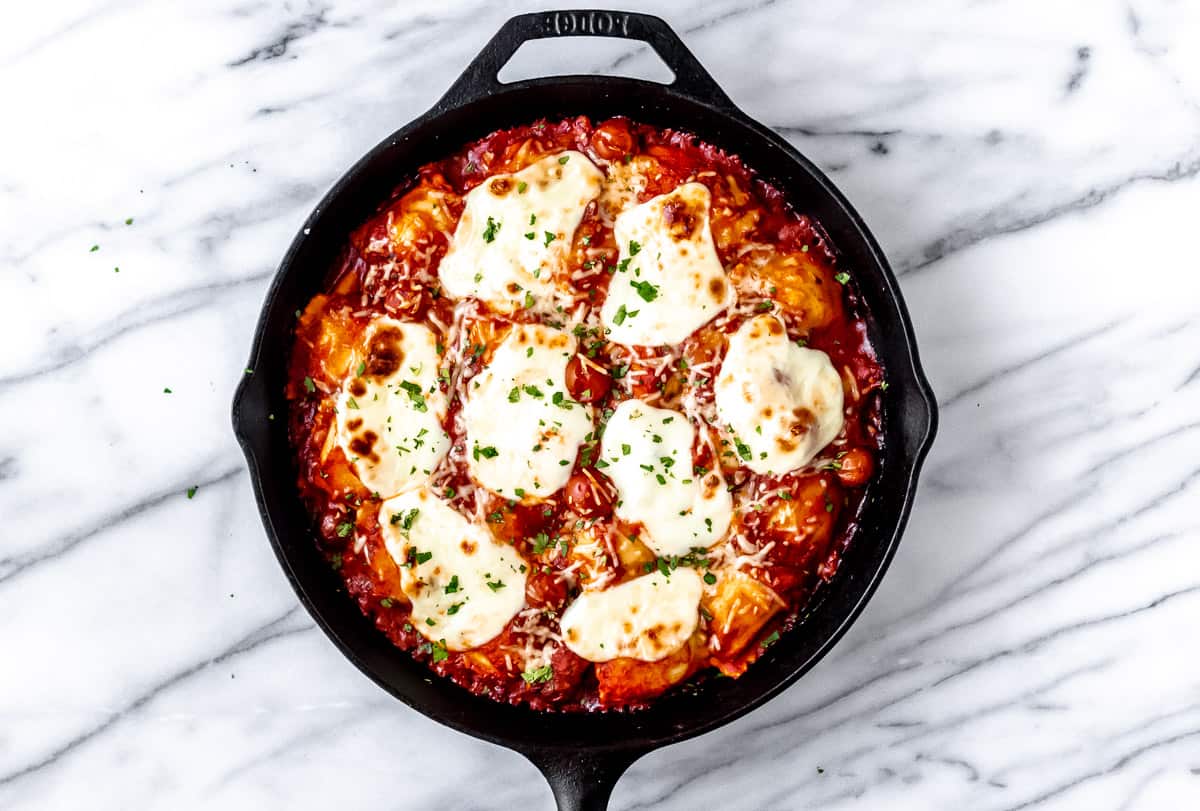 Baked ravioli casserole with fresh parsley in a cast iron skillet on a marble backdrop
