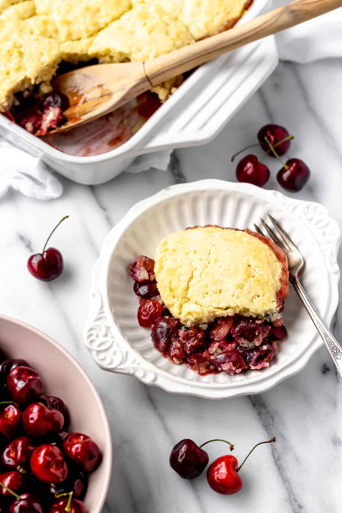 A serving of cherry cobbler in a white bowl with a bowl of cherries partially showing in the front, a baking dish of cobbler in the background and several cherries scattered around