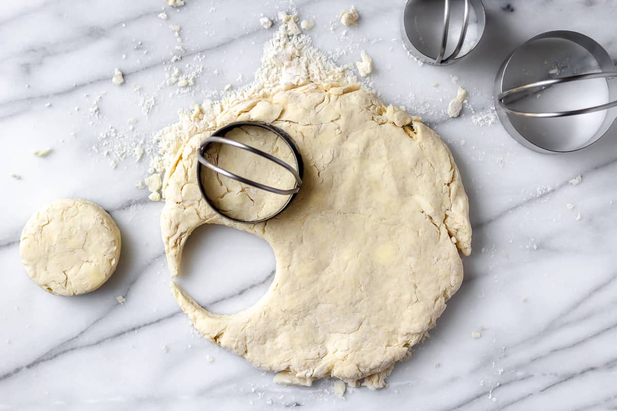 Biscuit dough and a one biscuit cut out with a biscuit cutter cutting out a second biscuit on a marble background