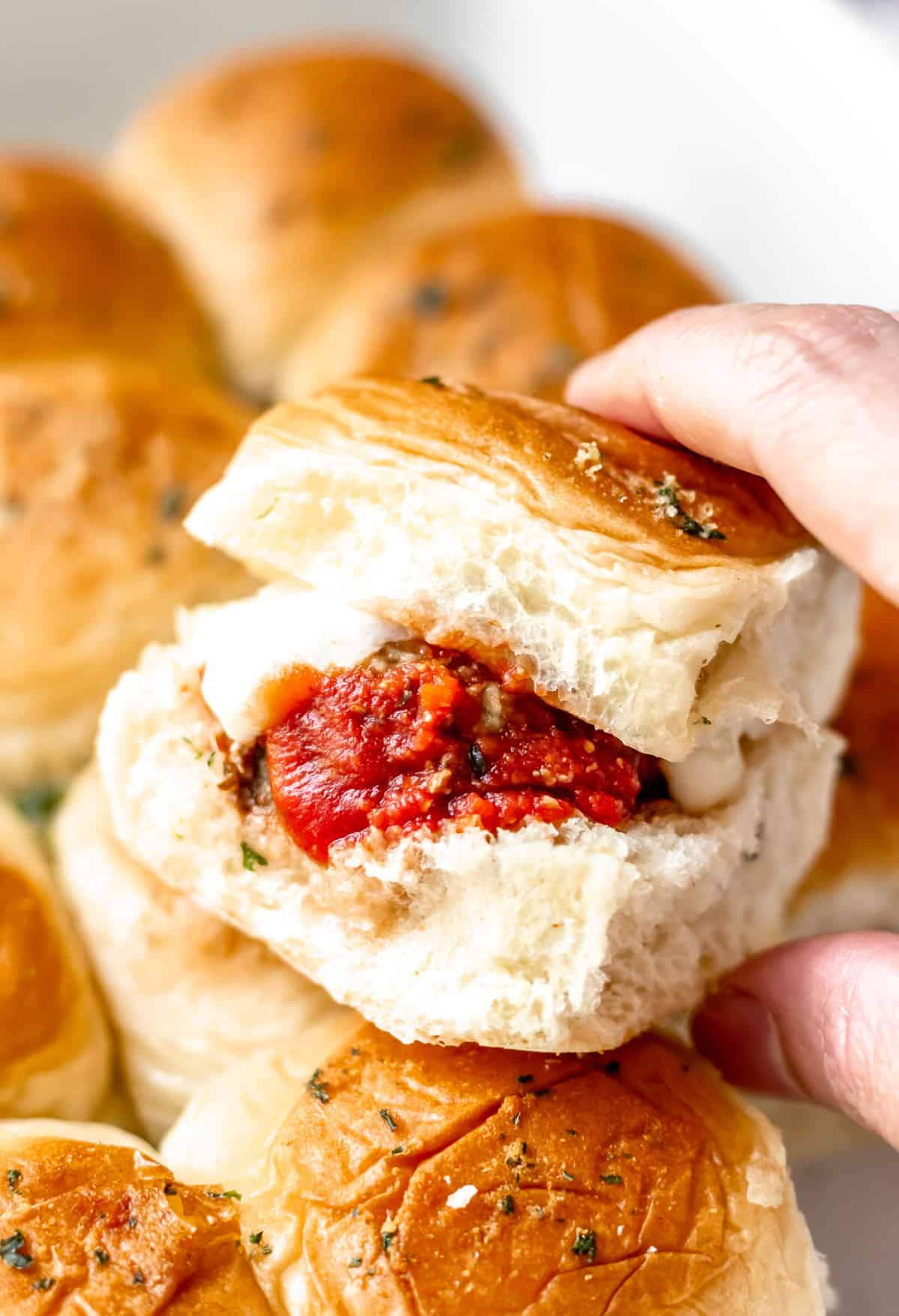 A hand holding up a meatball slider in front of the rest of the sliders still in the casserole dish