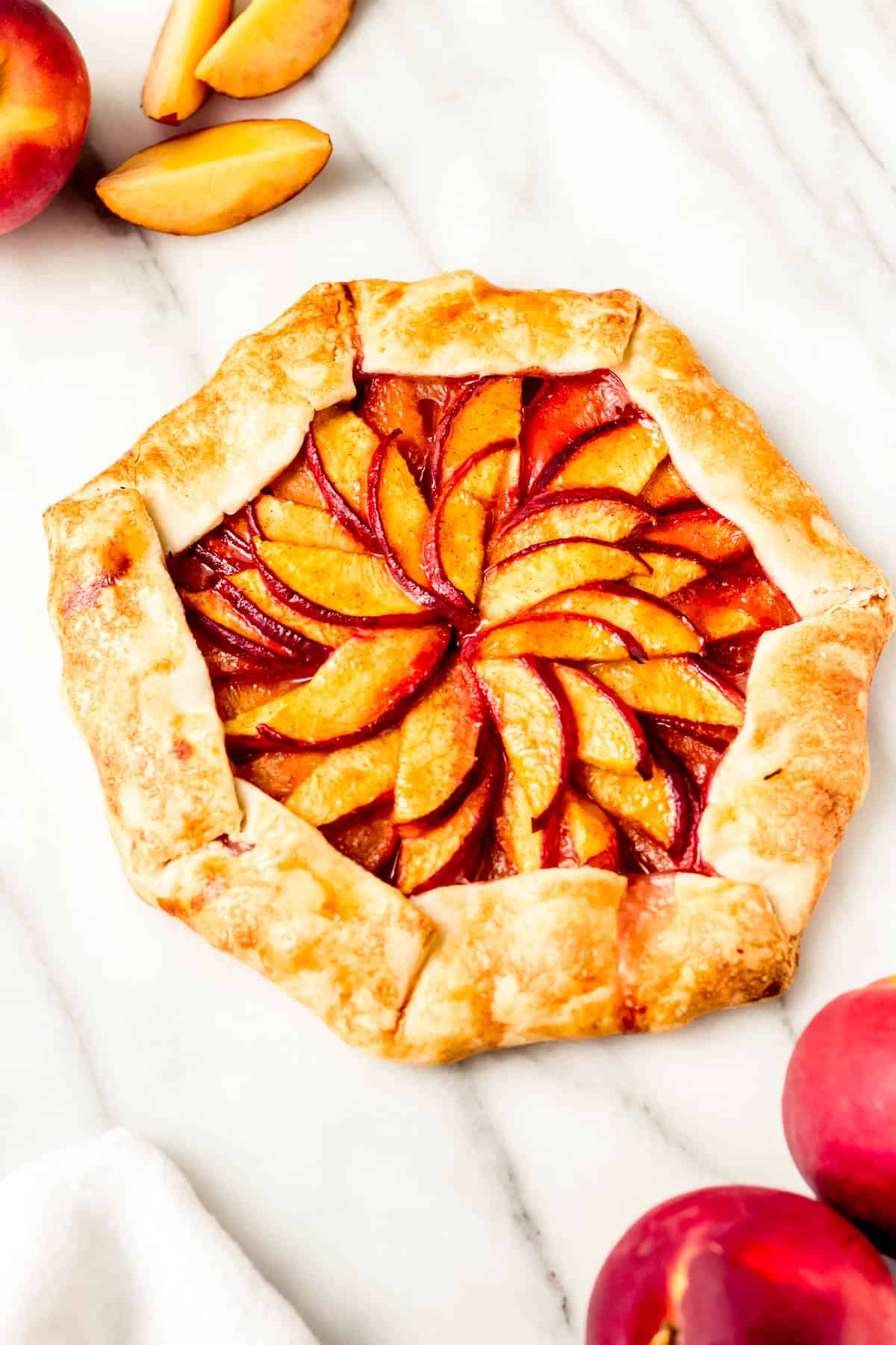 45 degree angle of a peach galette with whole and sliced peaches around it on a marble background