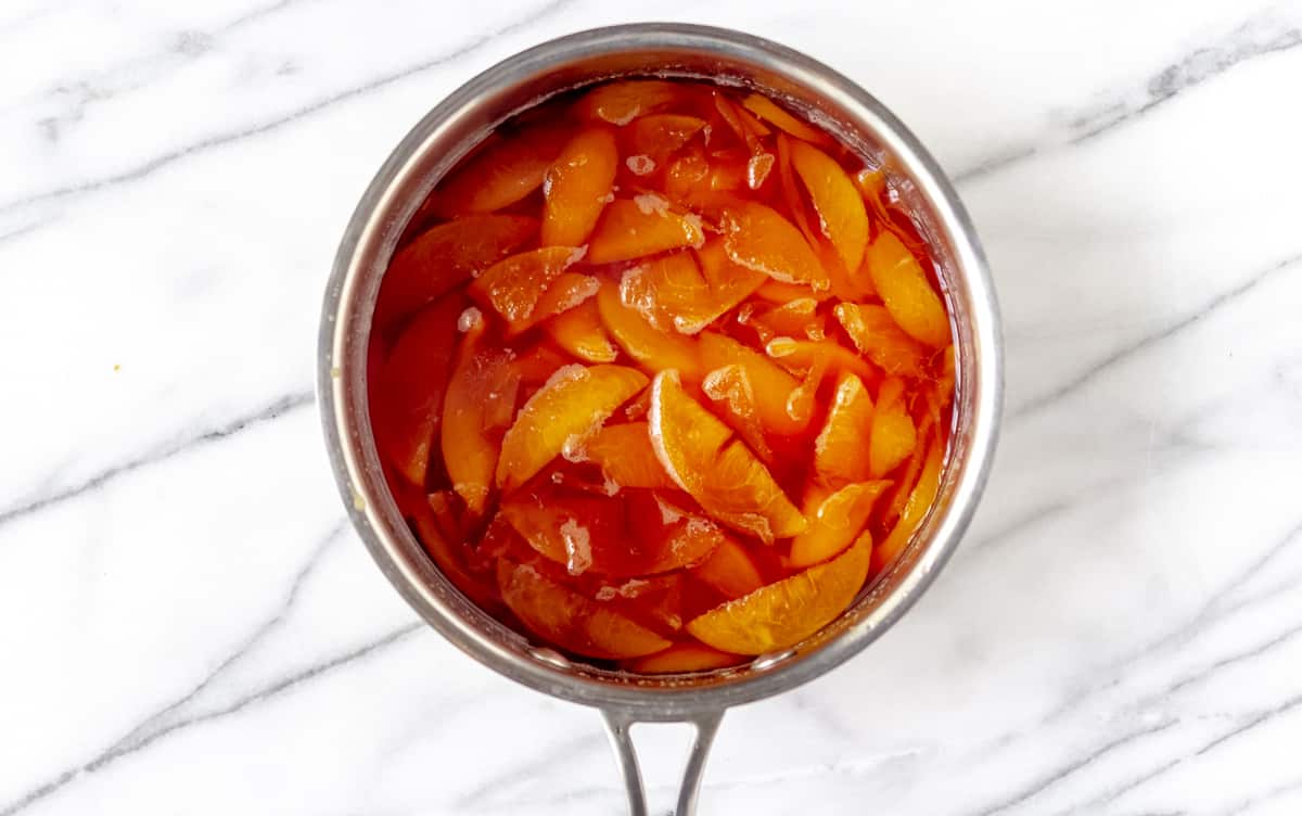 Cooked peaches in syrup in a sauce pot over a marble background