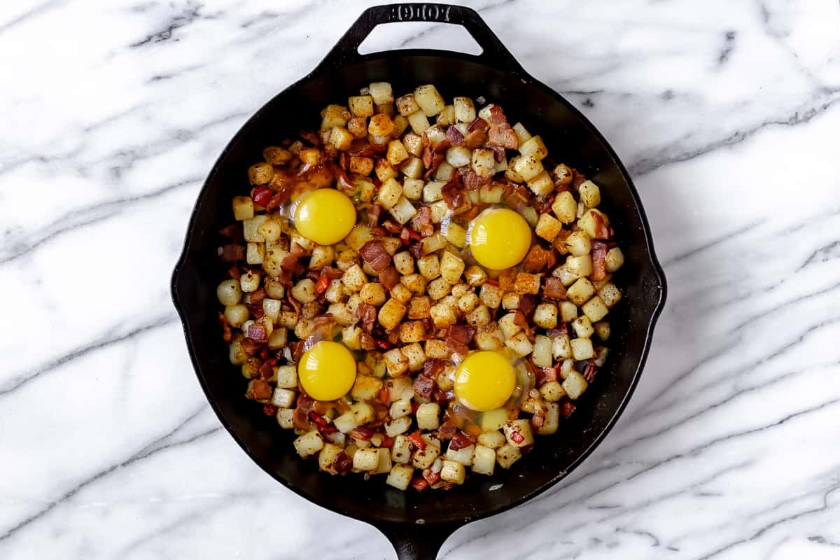 Potato hash with 4 eggs cracked into wells in a cast iron skillet before cooking the eggs