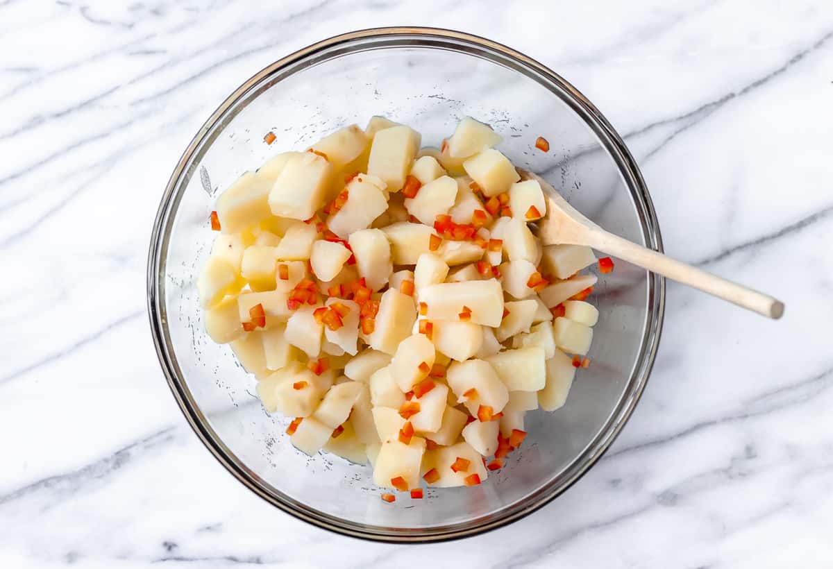 A large glass bowl with potatoes and red peppers in it and a wood spoon over a marble background