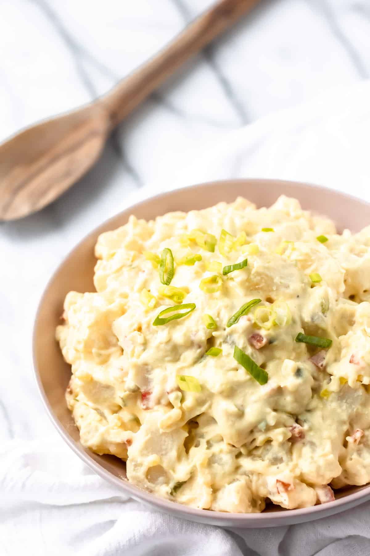 A pink serving bowl filled with amish potato salad and a wood spoon in the background