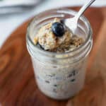 Blueberry overnight oats in a jar with a spoonful being held over it on a wood board.
