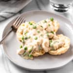 Creamed Chicken Over Biscuits on a white plate on a marble background with a fork, glass and towel partially showing