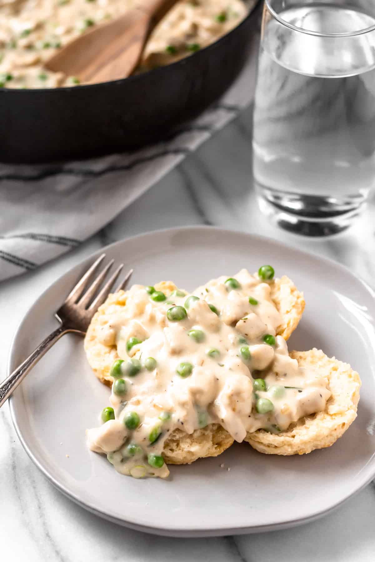 Creamed Chicken over biscuits on a plate with part of the skillet showing in the background and a glass of water, fork and towel 