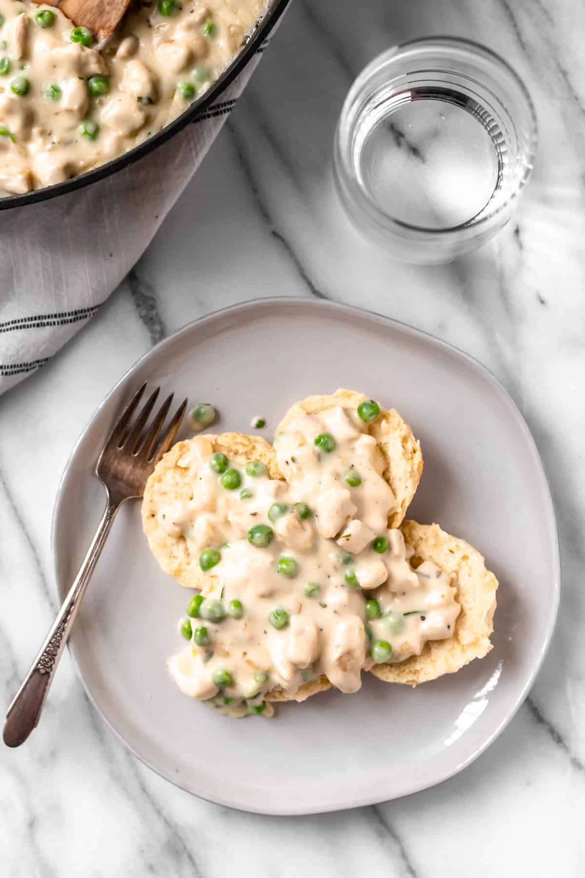 Overhead of creamed chicken over biscuits on a plate with part of the skillet, a fork, a glass and a towel around it