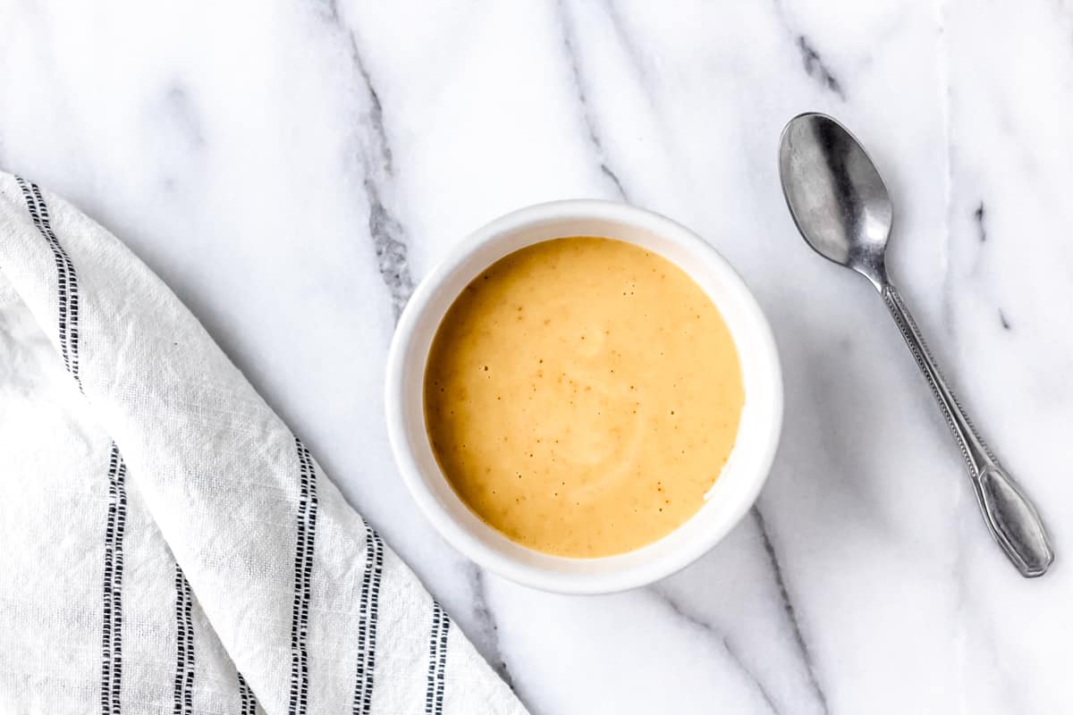 overhead of honey mustard sauce in a small white bowl with a spoon on one side and a part of a striped towel on the other