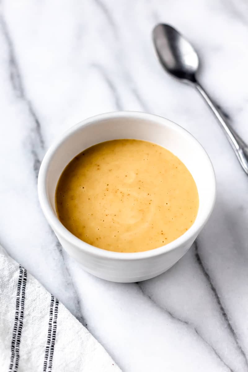 Close up of honey mustard dipping sauce in a small white bowl with a spoon off to the side and part of a towel showing on the other side