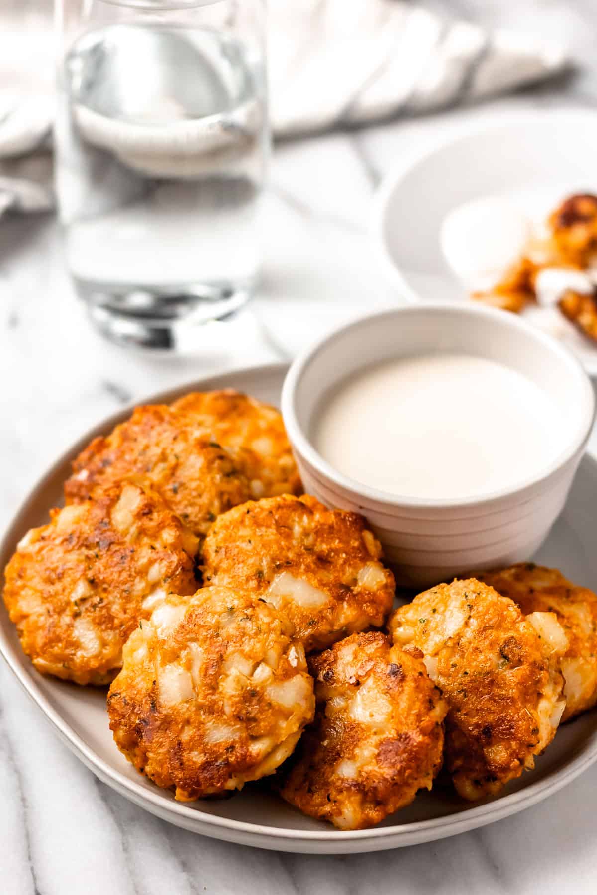 A plate of cod cakes with a white bowl of aioli and a second plate, glass of water and towel in the background.