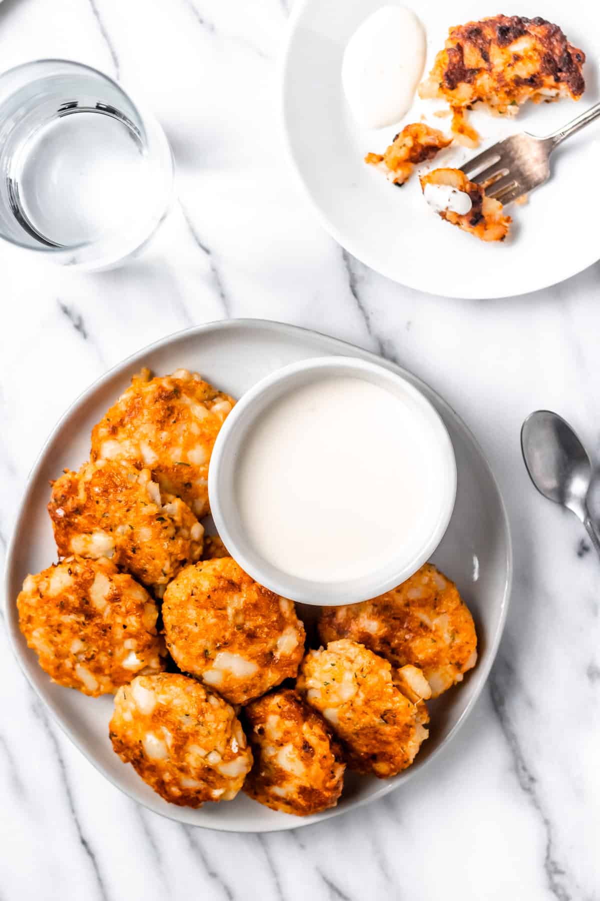 Overhead of cod cakes on a larger white plate with a white bowl of aioli, a second plate with a cut up cod cake and fork, a glass of water and a spoon all over a marble background.