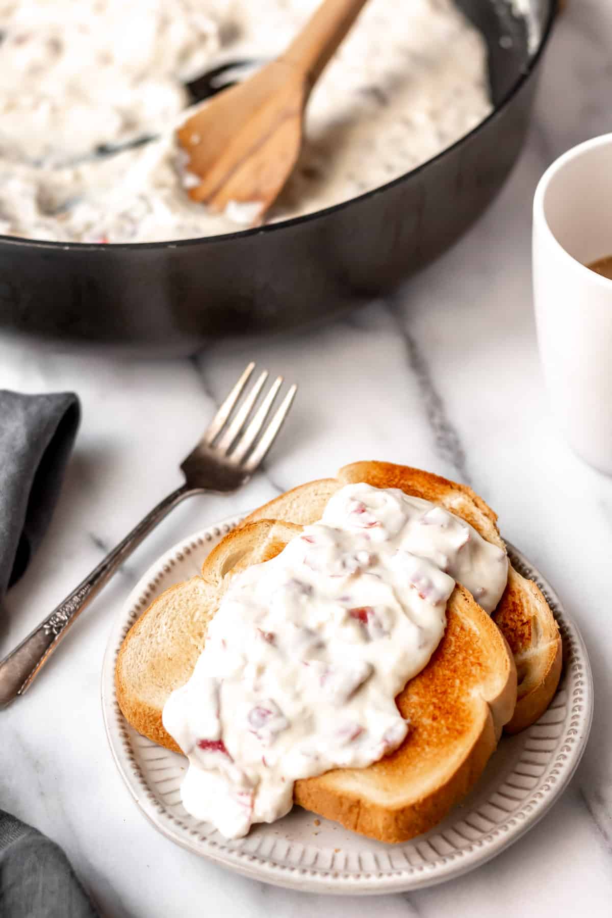 2 slices of Creamed Chipped Beef on toast on a white plate with a fork, gray towel, mug and skillet in the background all over a marble background.