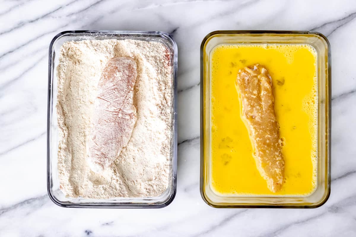 A chicken strip in a bowl of flour and a chicken strip in a bowl of whisked eggs over a marble background.