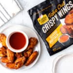 A plate of chicken wings with a bowl of Buffalo sauce and a package of Foster Farms Take Out Crispy Wings.