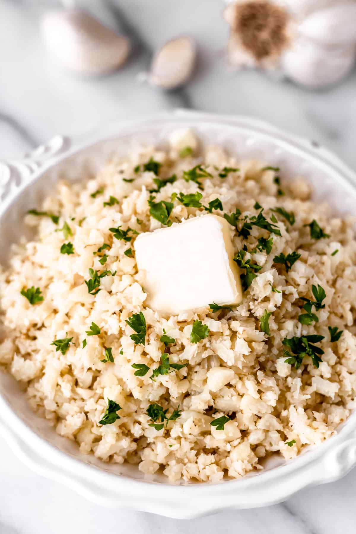 Close up of garlic cauliflower rice in a white bowl with garlic cloves in the background