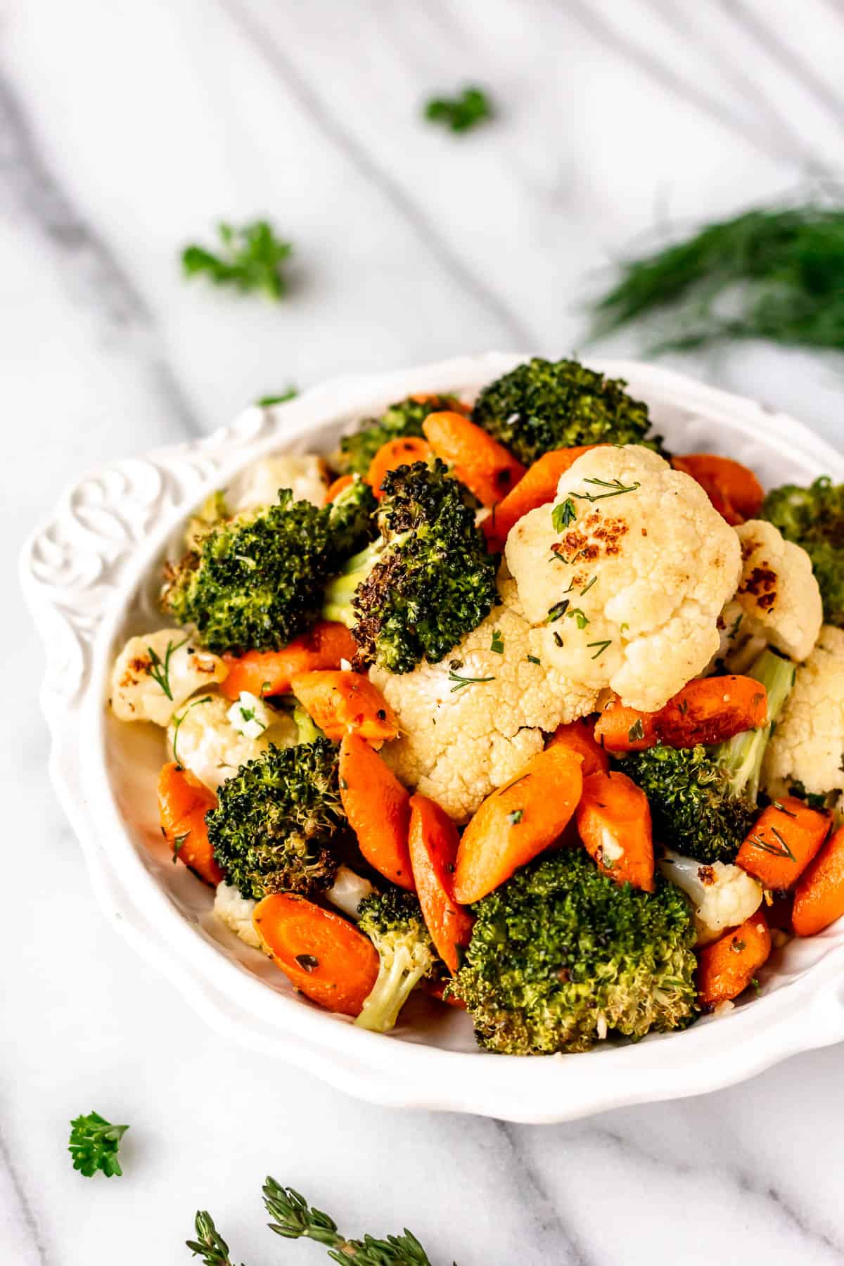 Angled view of a white bowl of garlic herb California blend vegetables with fresh herbs around it.