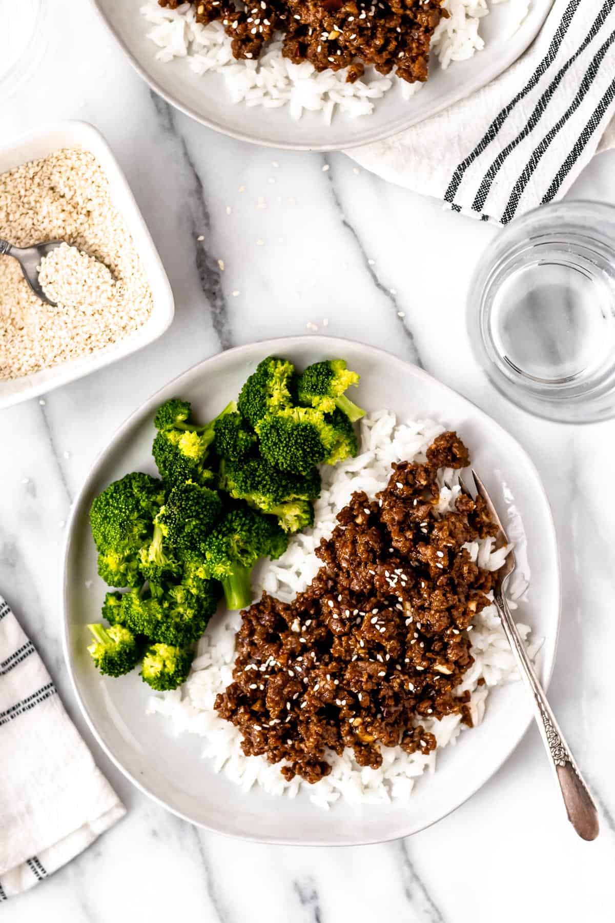 Overhead of a plate of Mongolian ground beef on rice with broccoli, a second plate partially showing and 2 towels, a glass of water and sesame seeds around the plates.