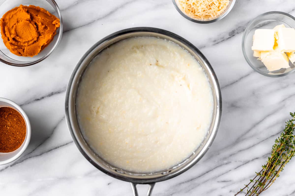 A silver pot of grits with other ingredients around it on a marble background.