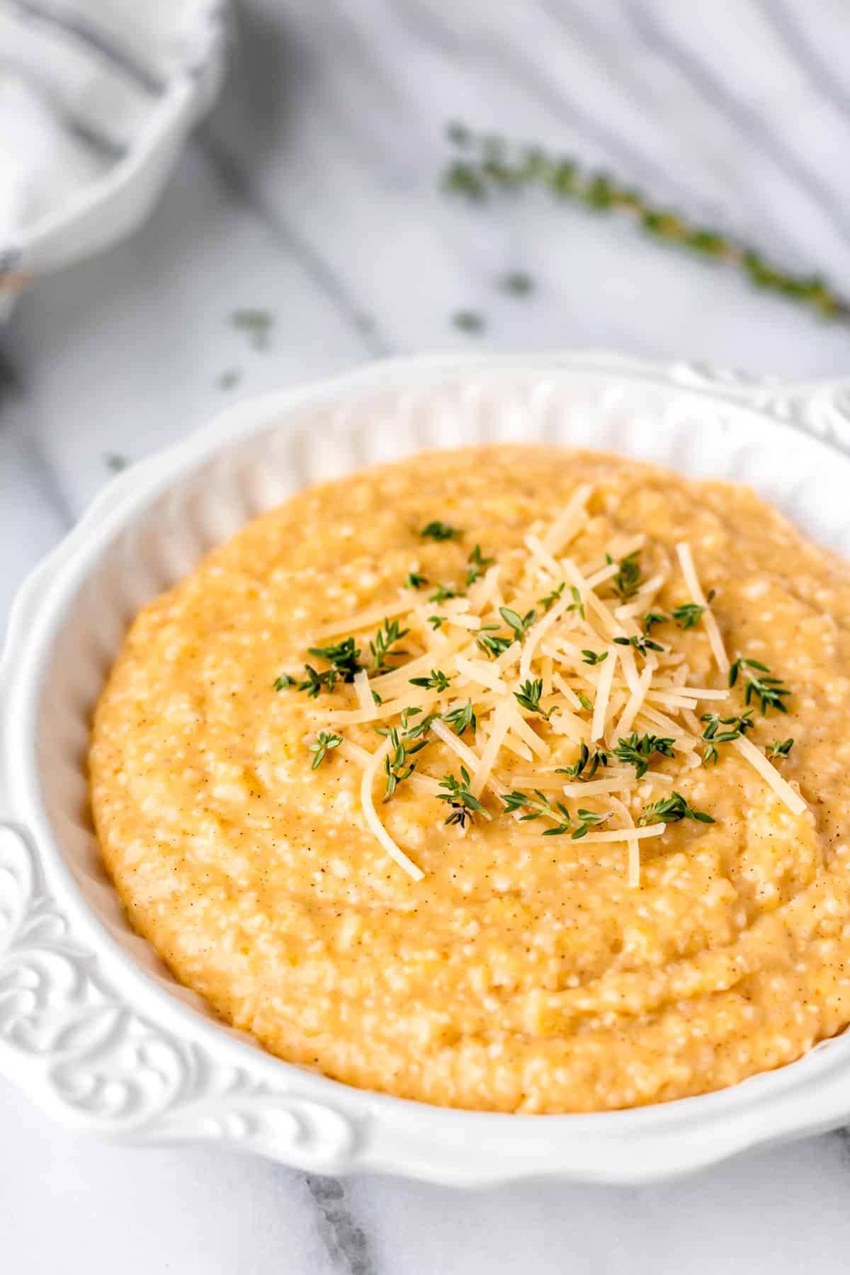 A white bowl of pumpkin grits with thyme and a towel in the background on a marble backdrop.