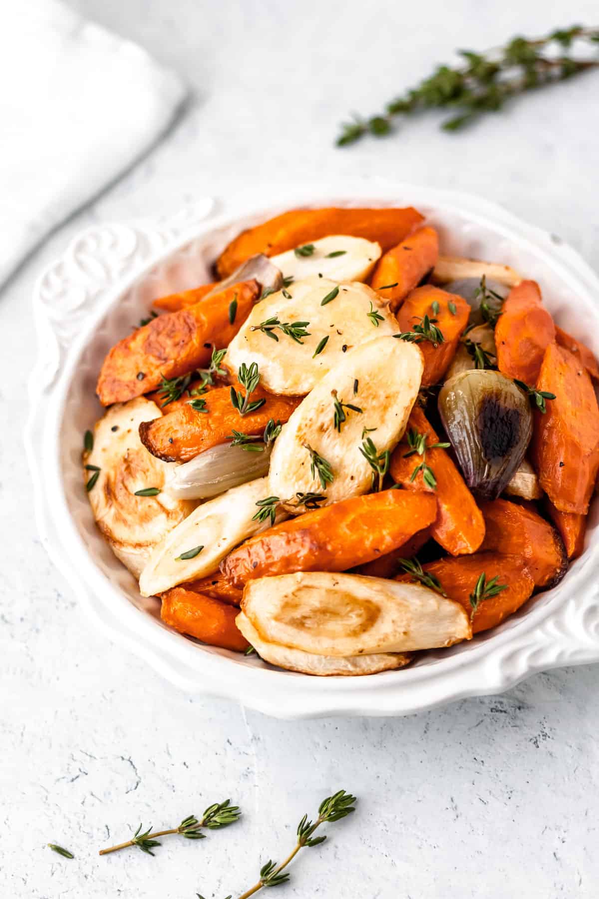 A white bowl filled with roasted carrots, parsnips, shallows and thyme with fresh sprigs of thyme around the bowl and a white towel in the background.
