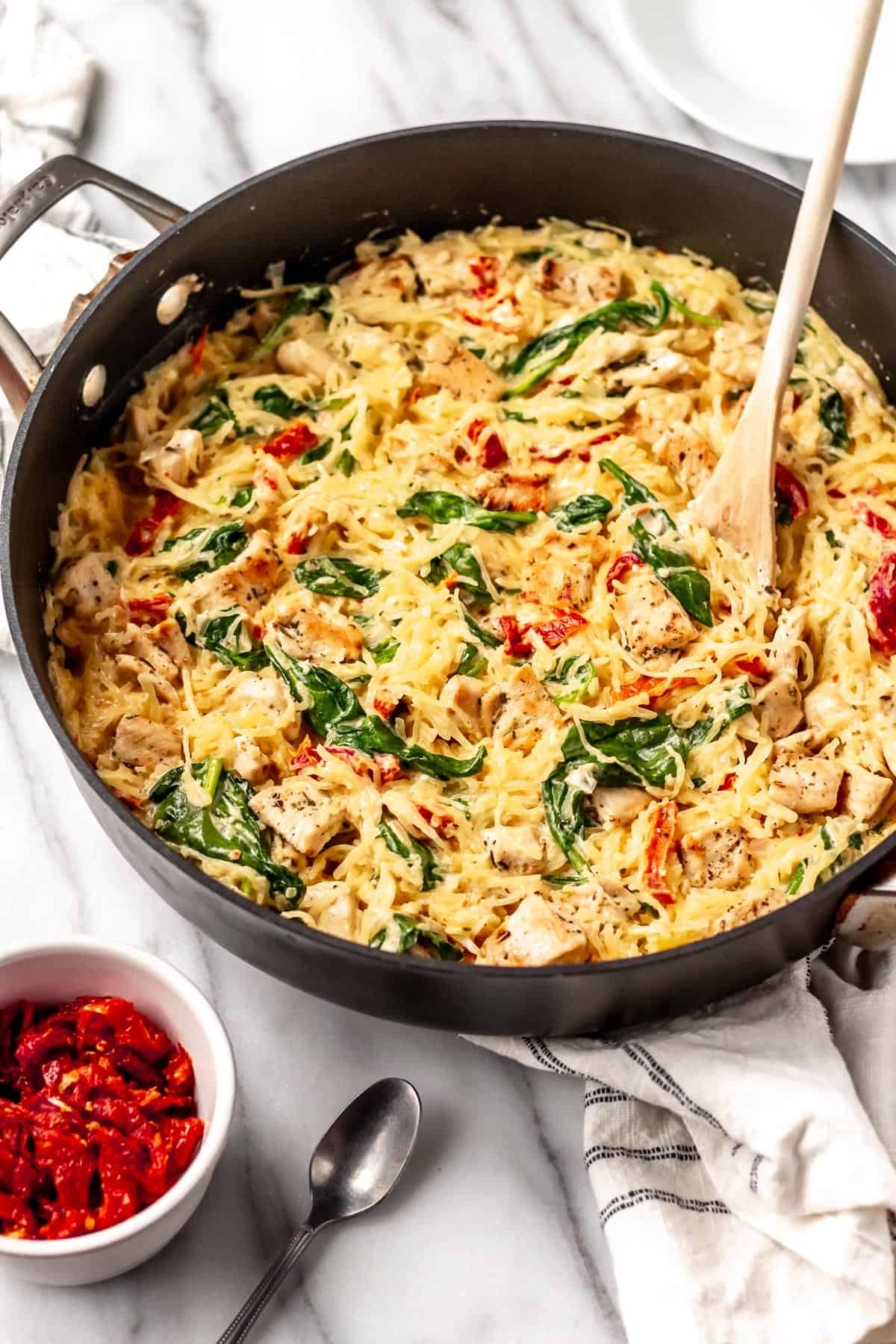 A black skillet with Tuscan chicken and spaghetti squash with a wood spoon in it. There is a small bowl of sun-dried tomatoes in a white bowl, a spoon and towel around it.