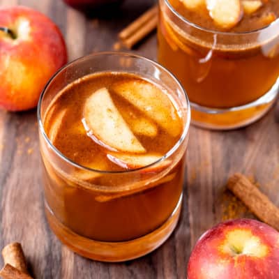 Apple brandy cider in a glass with apple slices with a second glass, apples and cinnamon sticks in the background.