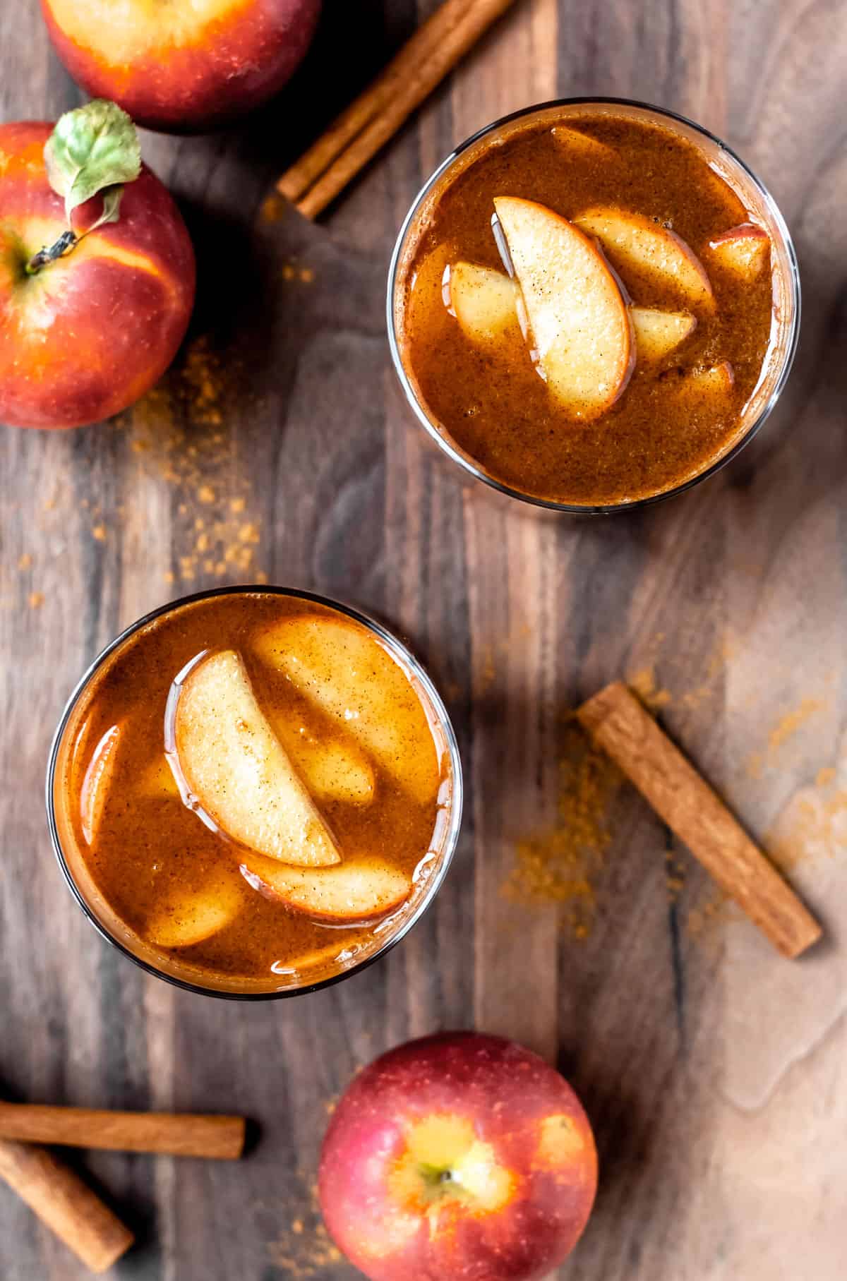 Overhead of 2 apple brandy cider drinks with apple slices on top and apples and cinnamon sticks around them on a wood background.