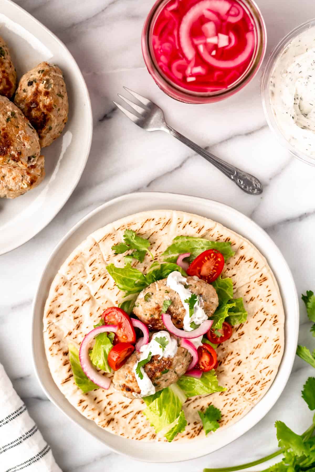 Overhead of pita with chicken kofta, pickled onions and lettuce with a plate of more kofta, jar of pickled onions, fork and towel around it.