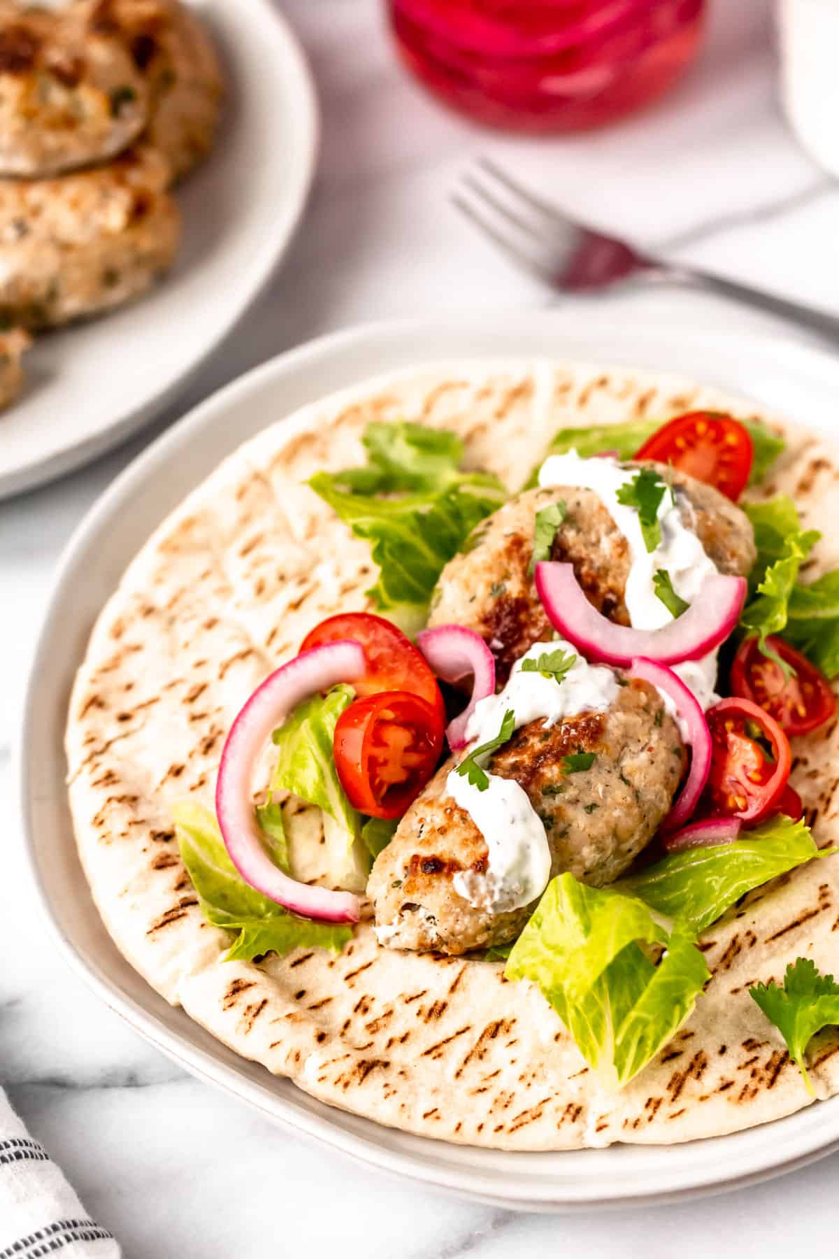 Close up of a plate with chicken kofta pita with a fork, second plate and jar of pickled onions partially showing in the background.