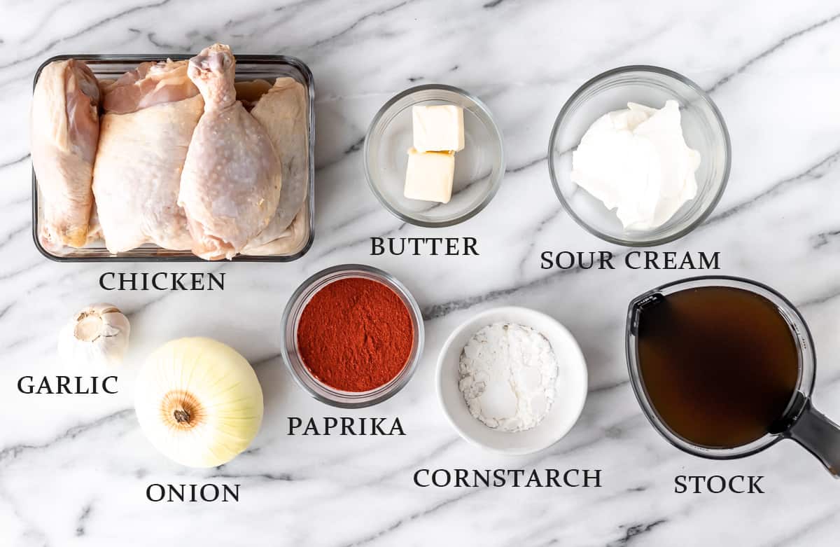 Ingredients needed to make chicken paprikash laid out on a marble backdrop with text overlay.