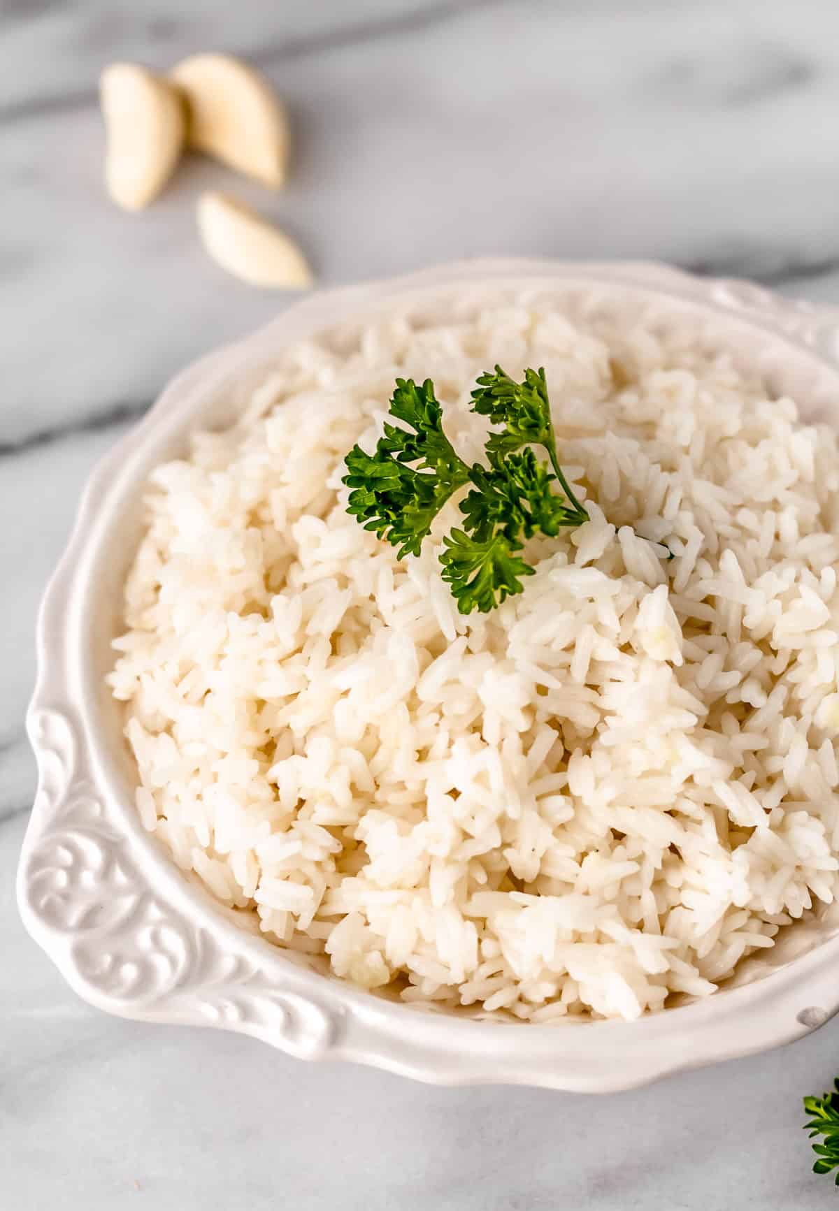 Close up of garlic rice in a white bowl with parsley on top and cloves of garlic in the background.