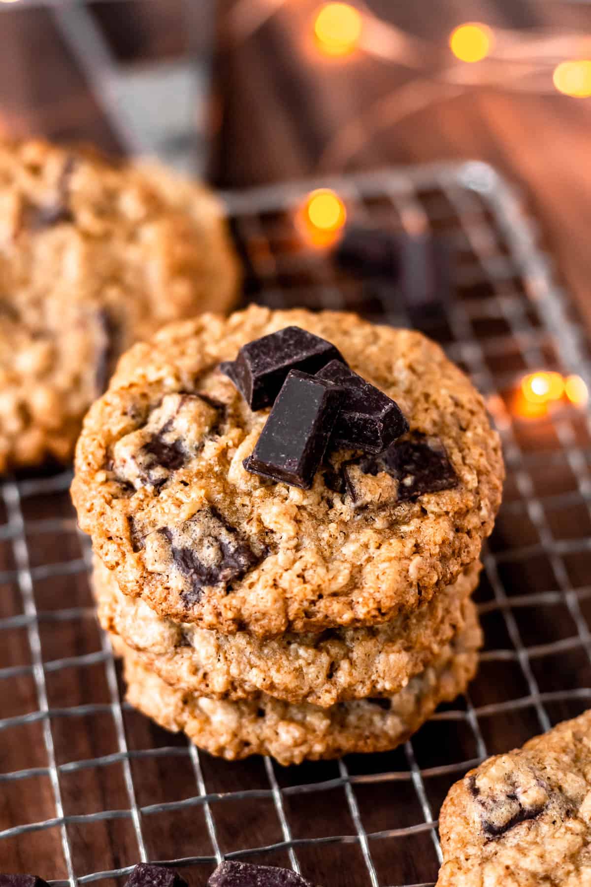 A stack of Oatmeal Chocolate Chunk Cookies with chocolate chunks on top over a cooking rack and wood table with lights in the background.
