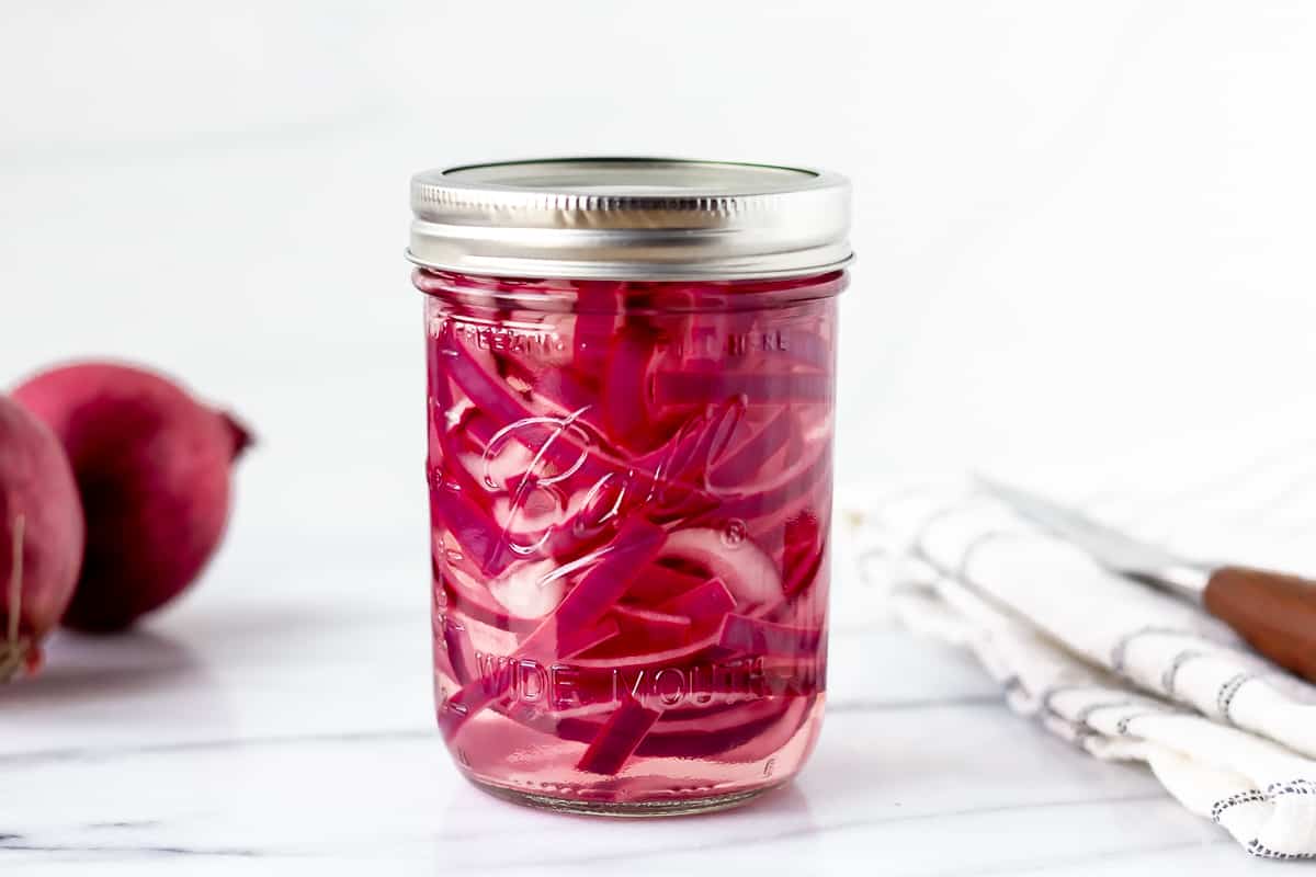 Pickled Red Onions in a canning jar with an onion and towel in the background.