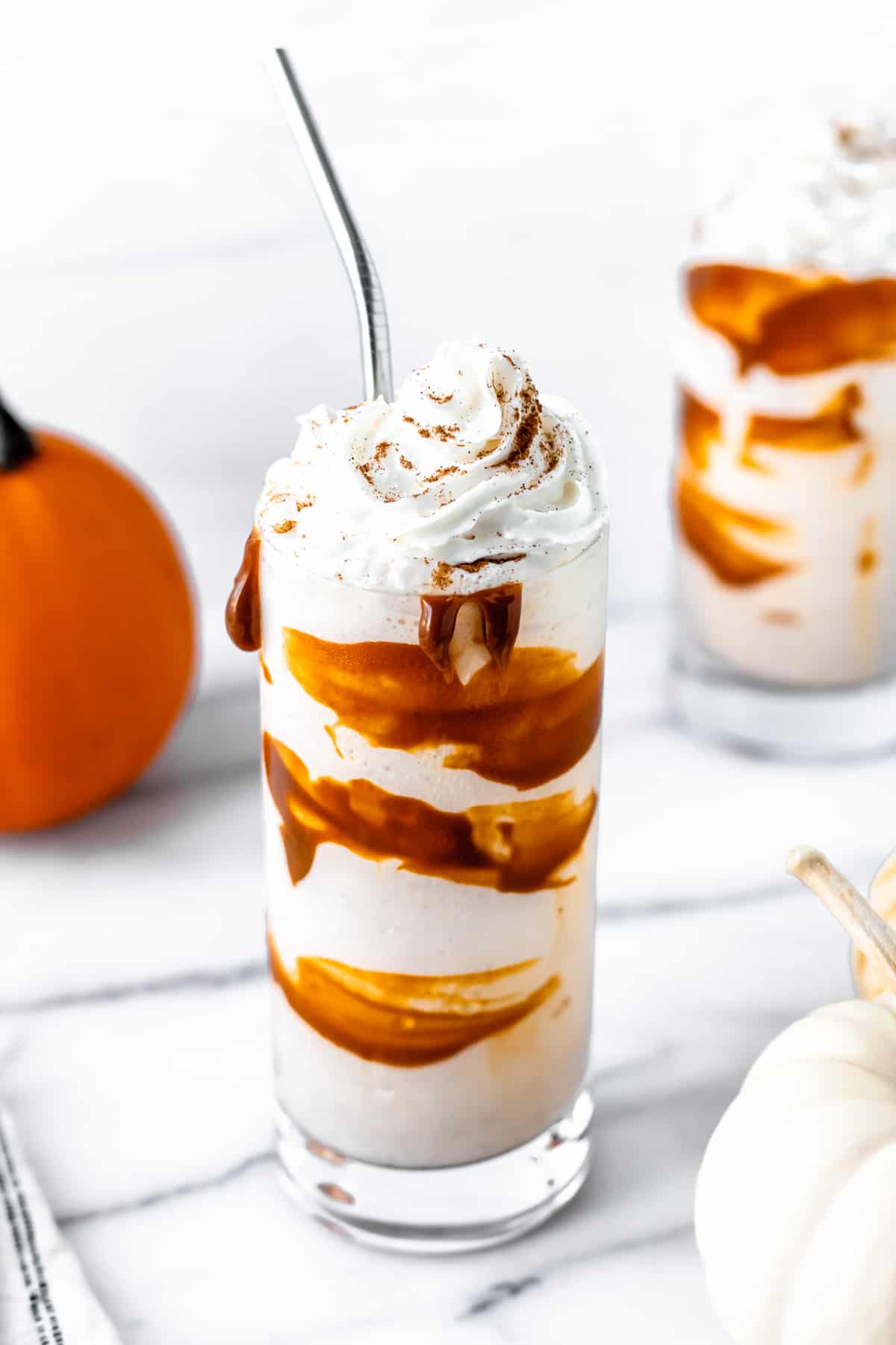 Slight overhead of a boozy pumpkin spice milkshake with a metal straw, second glass and pumpkins around it.
