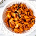 Close up of diced, roasted honeynut squash in a white bowl with fresh thyme garnish.