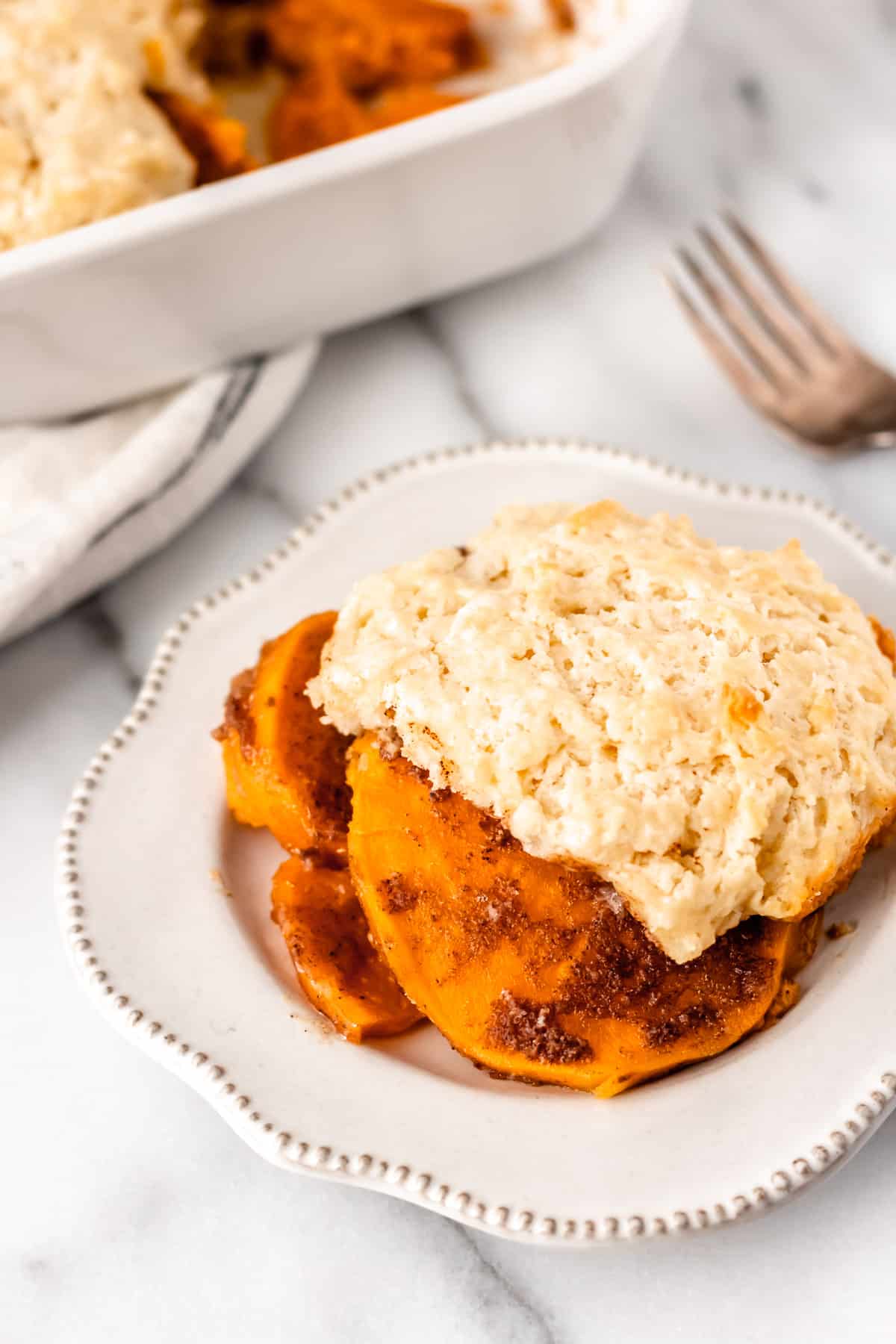 A serving of sweet potato cobbler on a small white plate with a fork and baking dish in the background.