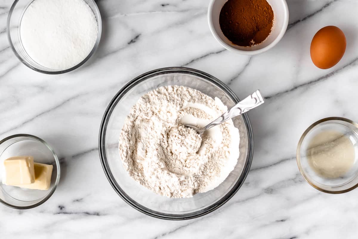 Dry ingredients for snickerdoodles in a glass bowl with a spoon and other ingredients around it on a marble table.