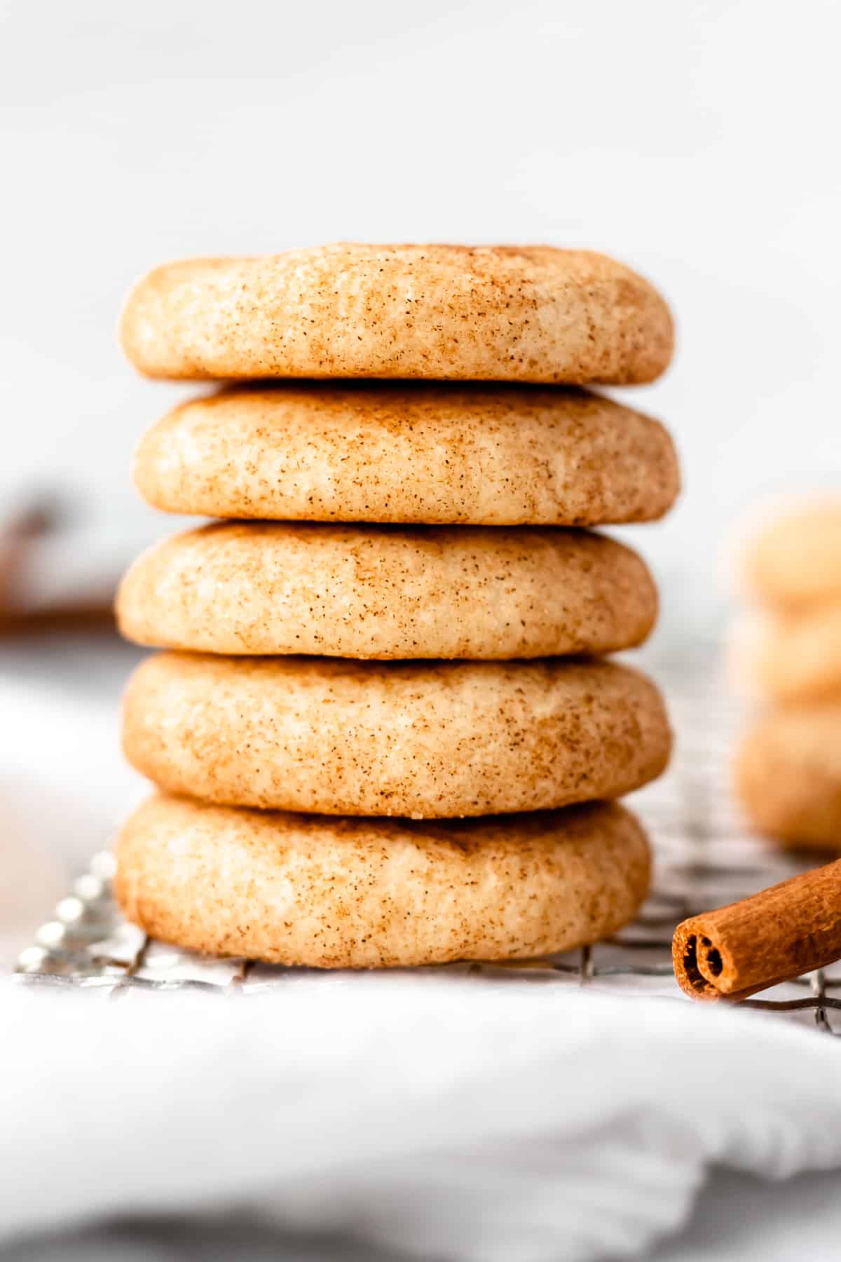 A stack of 5 snickerdoodle cookies on a cooling rack with cinnamon sticks and more cookies in the background.