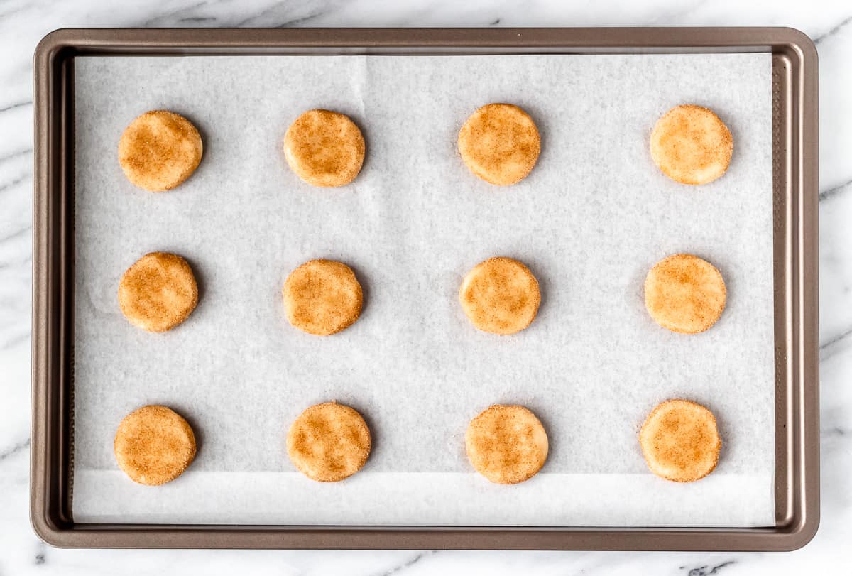 Snickerdoodle cookie dough balls flattened on a parchment paper lined baking sheet.