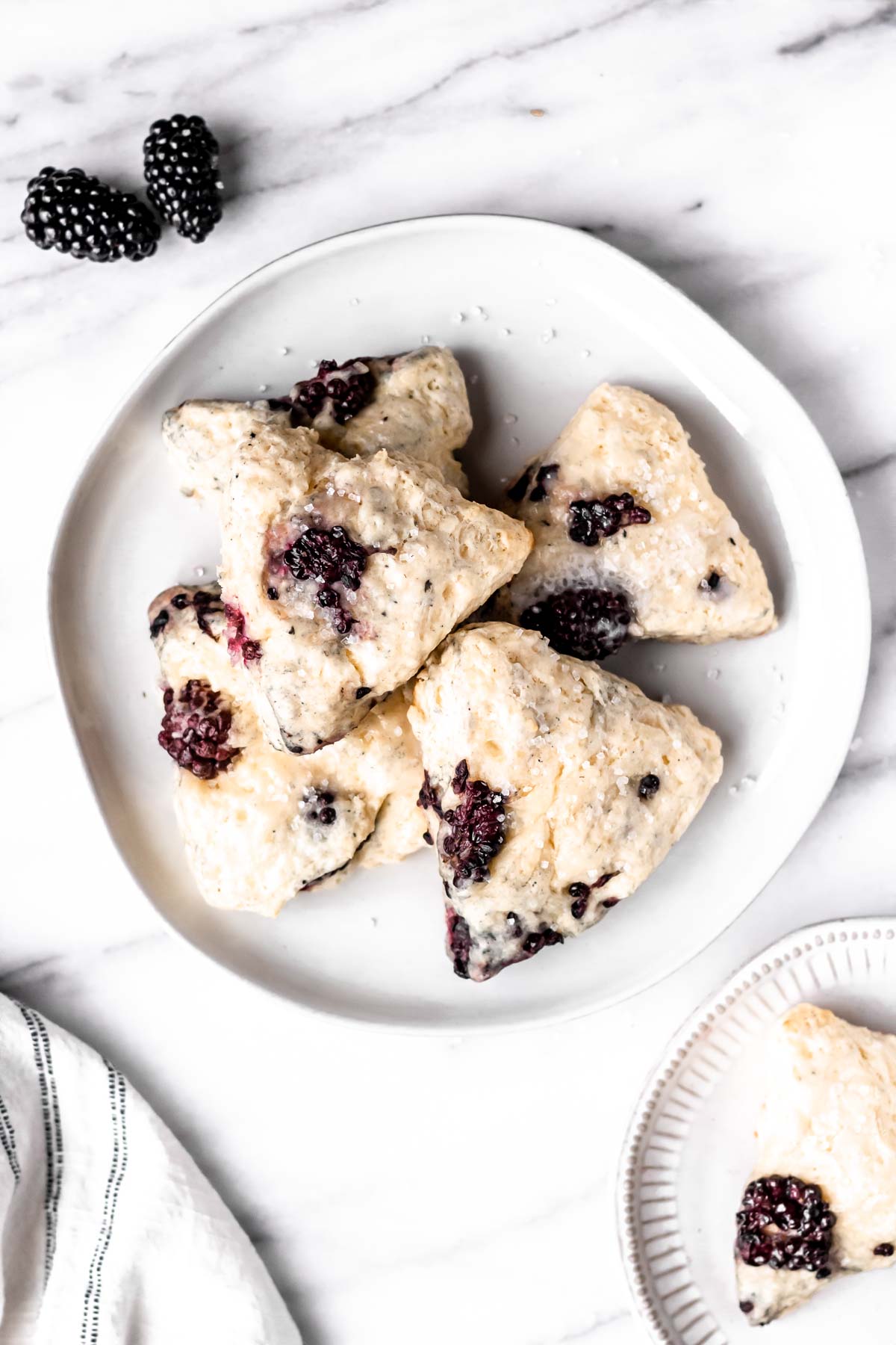 Overhead view of blackberry scones on a plate with a single one on a smaller plate, a white towel and extra blackberries.