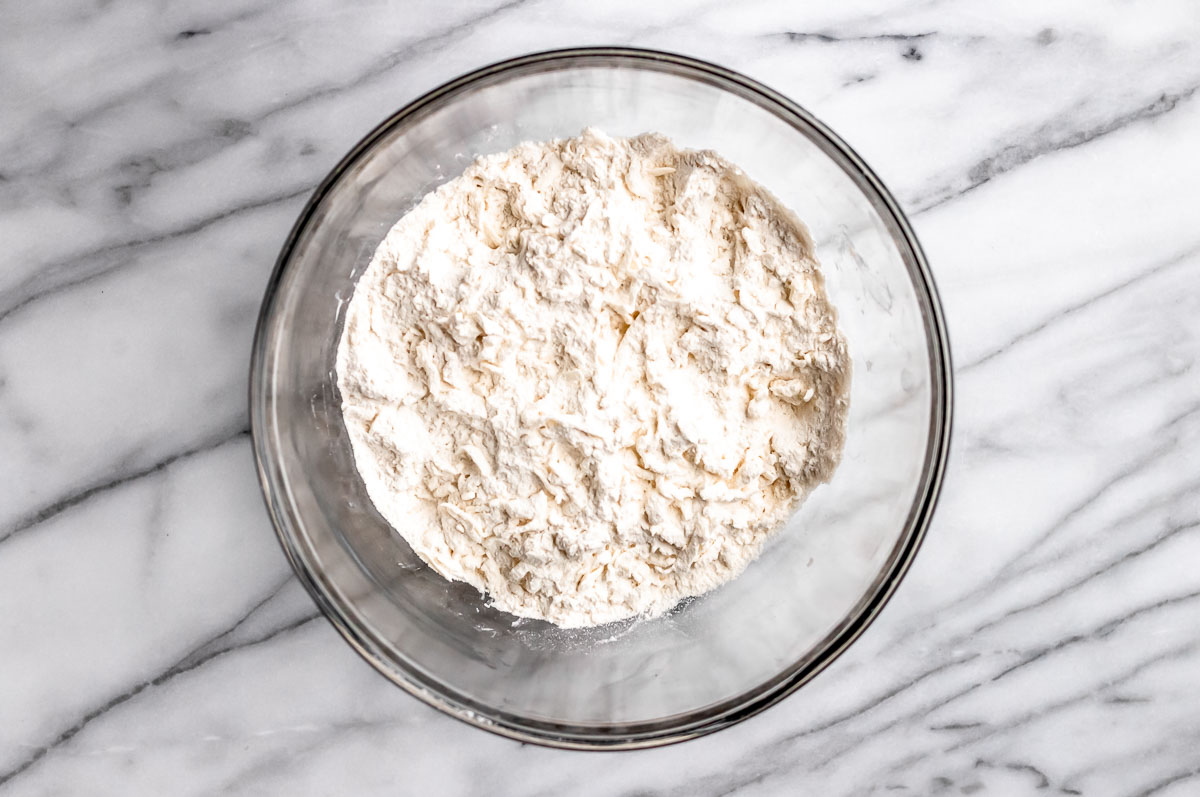 Dry ingredients with pieces of butter in a glass bowl on a marble background.