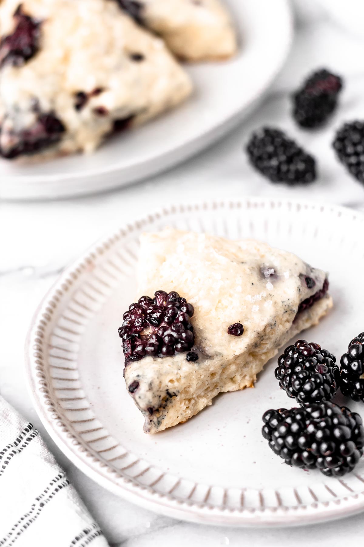 Close up of a blackberry scone on a small white plate with extra blackberries and a plate of scones in the background.