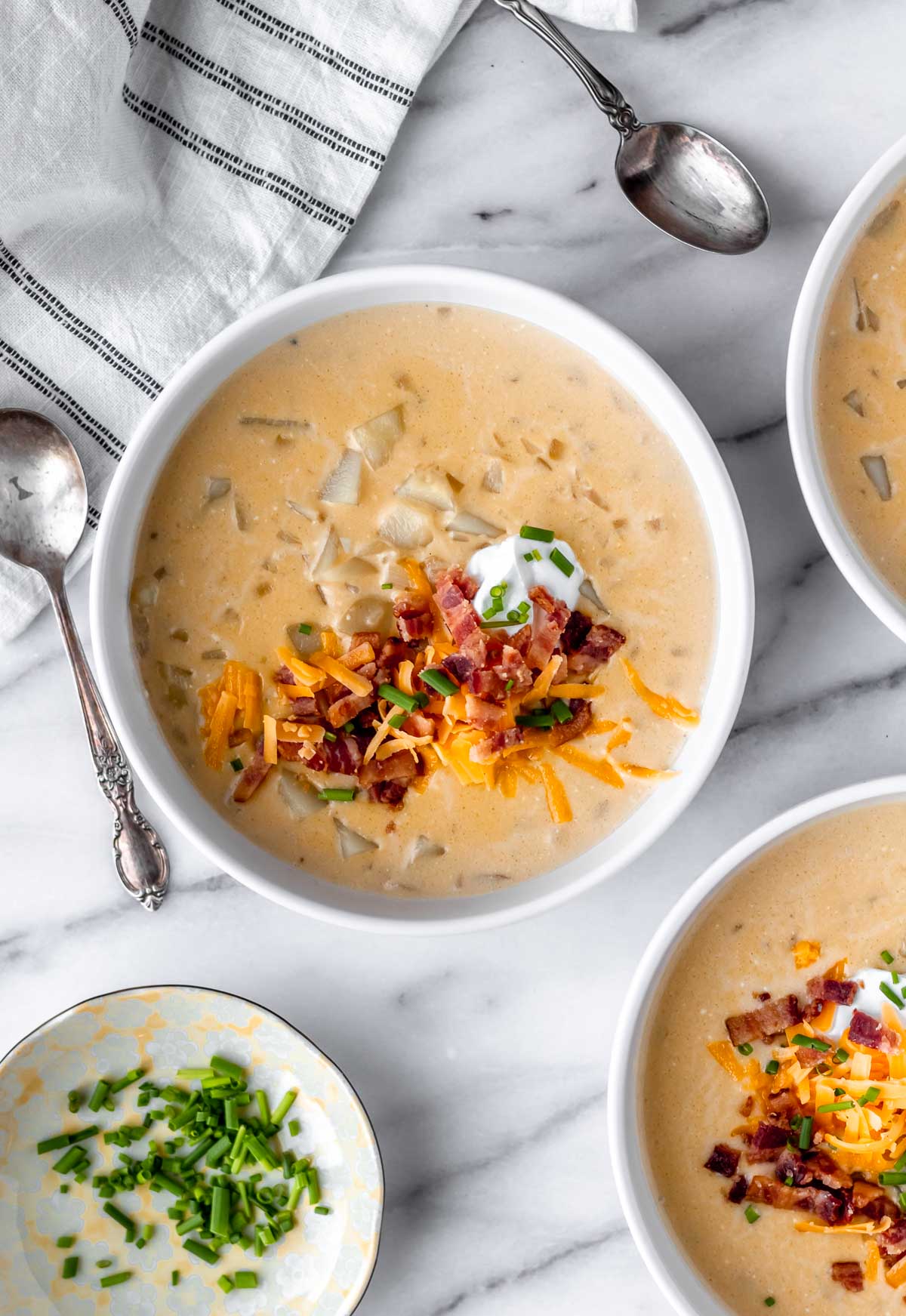 Overhead view of three bowls of loaded potato soup, a mini plate of chives, two spoons and a striped towel.