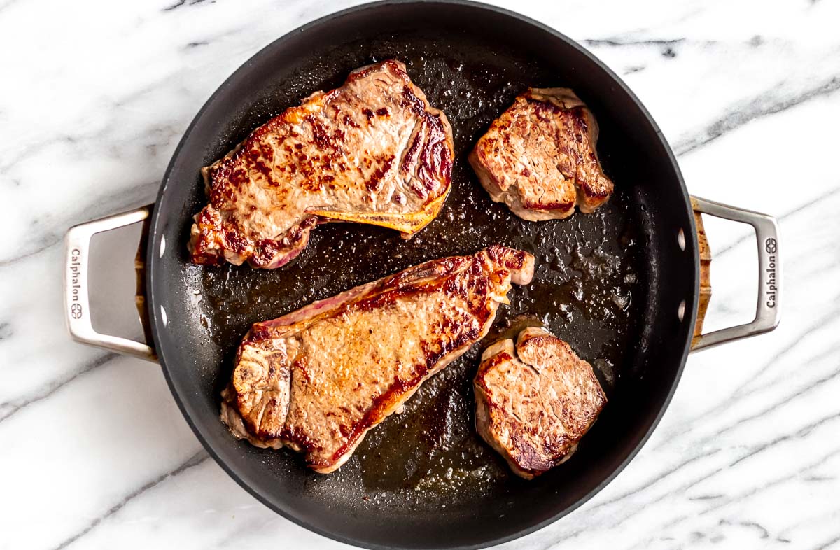 Four steaks cooking in a skillet.