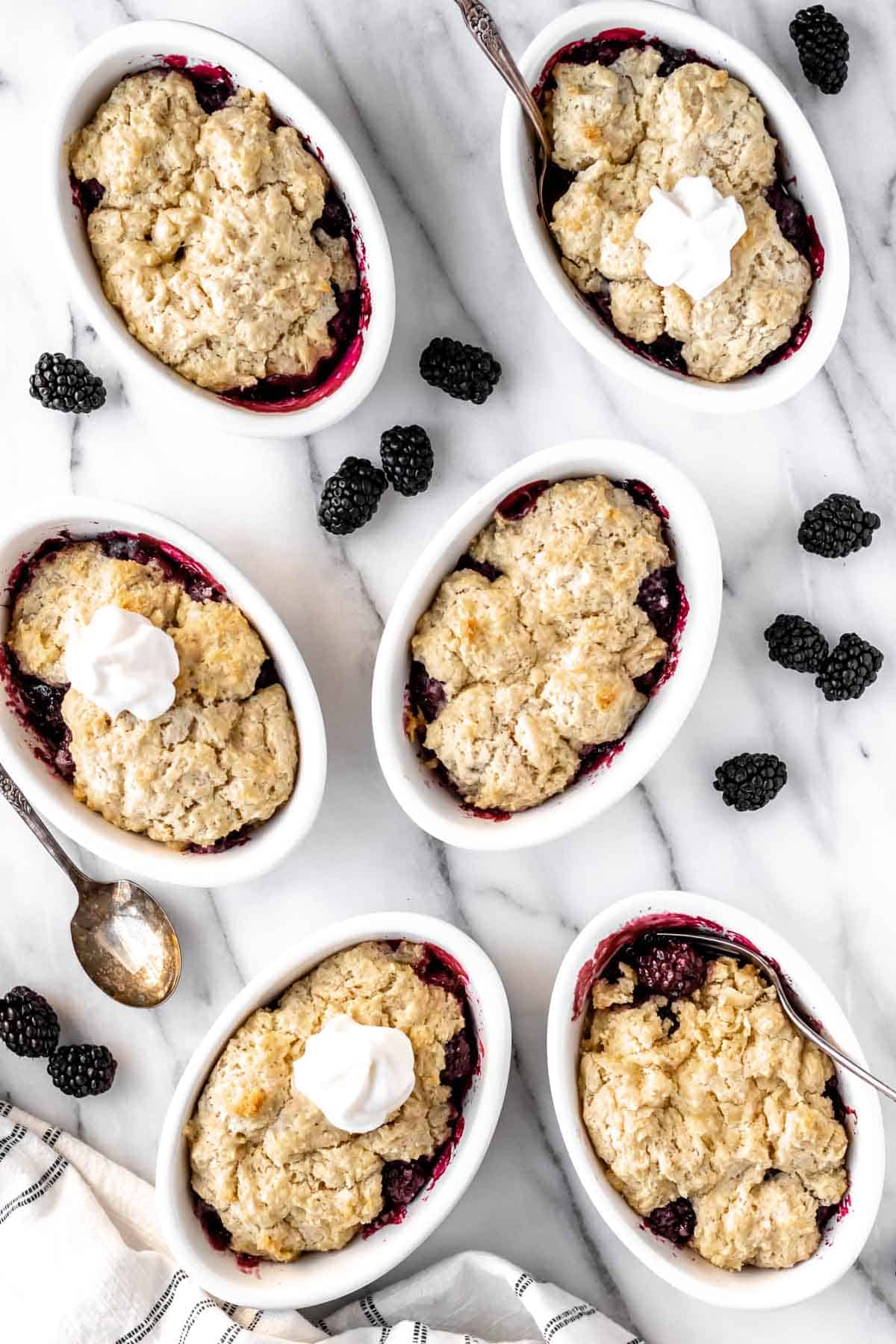 Six mini blackberry cobblers on a marble background with blackberries around them and forks in 2 of them.
