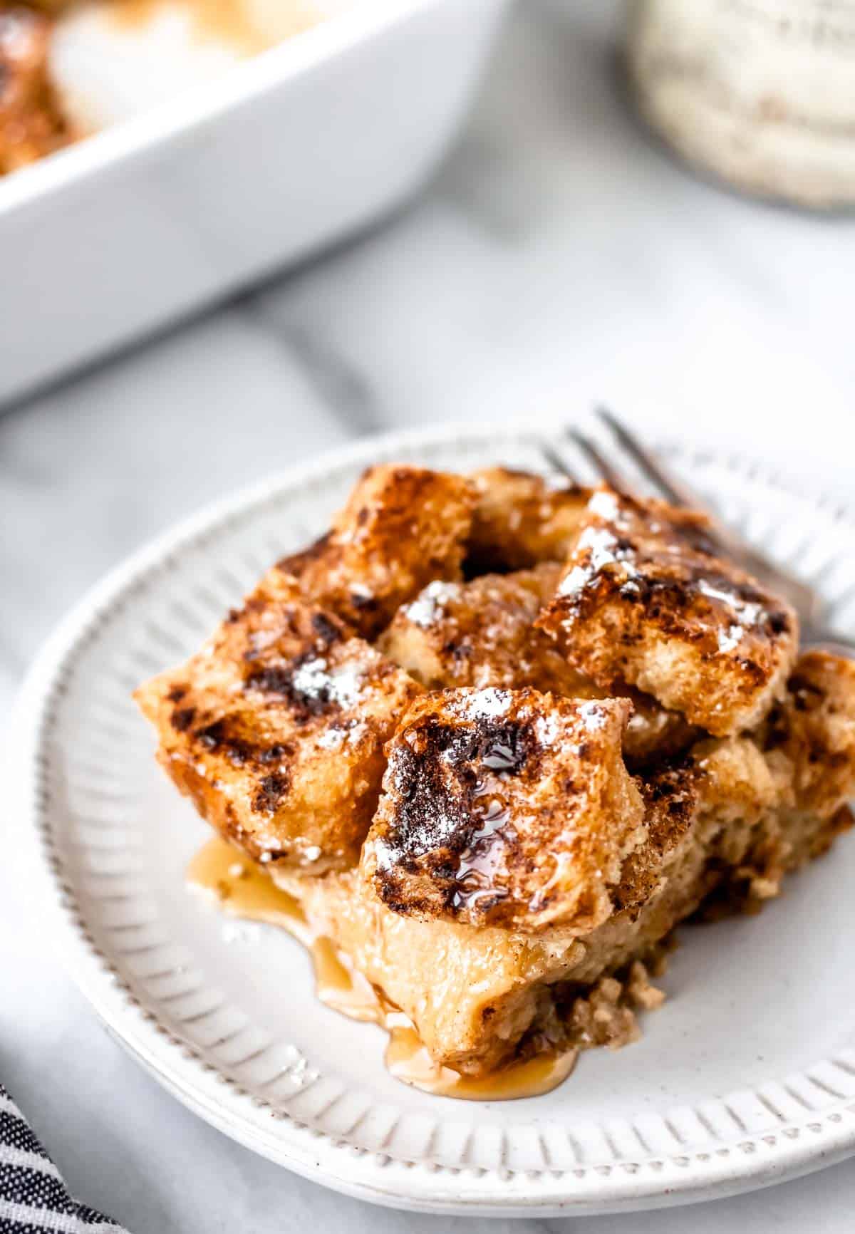 A serving of French Toast Casserole topped with maple syrup and powdered sugar on a small plate with a fork and part of the baking dish in the background.